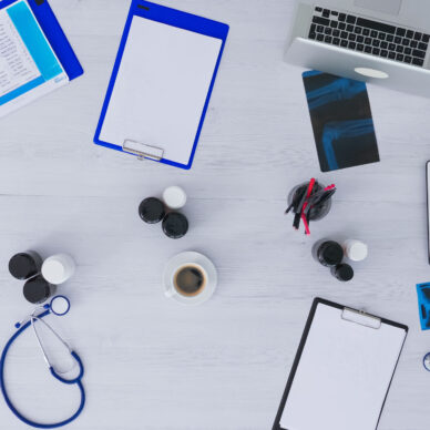 Top view of medical practitioner checking appointments using tablet, tacking notes on clipboard sitting on clinic desk with x-ray and medical equipment all around. Copy space, flat lay concept.